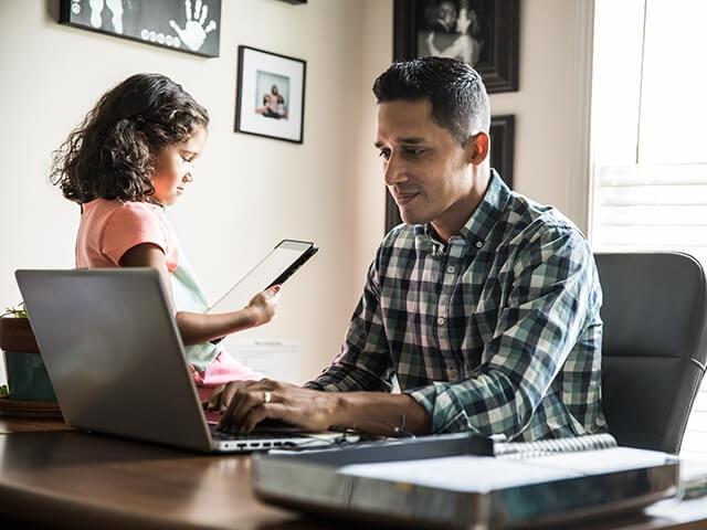 A father works at a desk on a laptop while daughter sits with him holding a tablet device