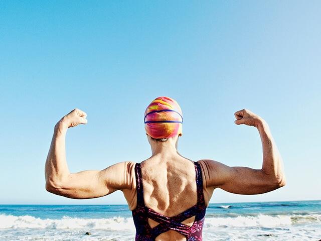 A female swimmer flexes biceps while standing on a beach facing ocean